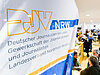 Ein Banner des DJV-NRW im Vordergrund; im Hintergrund mehrere Messestände und viele Menschen. –
