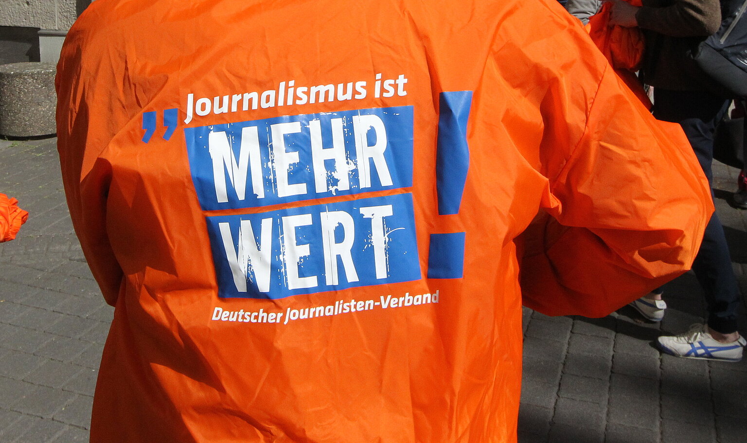 Ein Mensch mit dem Rücken zur Kamera in einer orangenen Jacke auf der steht: " Journalismus ist mehr wert!" –