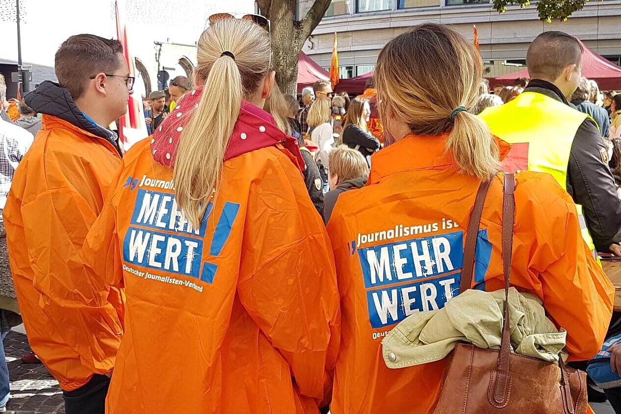 Eine Menge an streikenden Menschen, im Vordergrund drei junge Menschen in orangenen Jacken des DJV. – Journalismus ist mehr Wert! Foto. Corinna Blümel