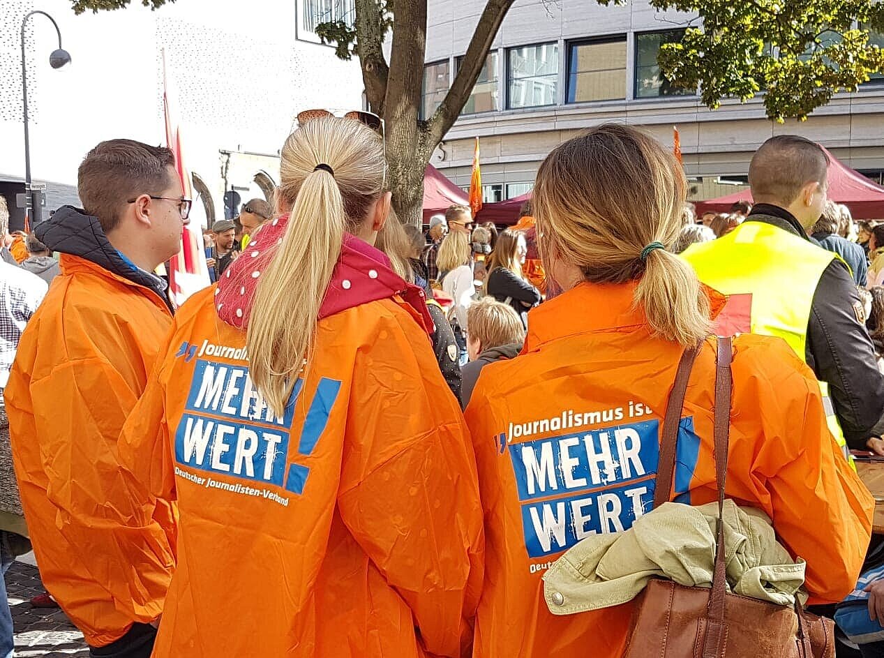 Eine Menge an streikenden Menschen, im Vordergrund drei junge Menschen in orangenen Jacken des DJV. –