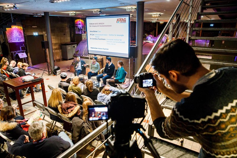 Ein Fotograf schießt ein Foto auf dem Branchentreff Journalistentag. Vier Personen sitzen auf dem Podium, die Zuhörer davor im Plenum. 