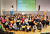 Ein Gruppenbild mit über 200 Menschen, die orangene Taschen und Zettel in die Luft halten. – 
