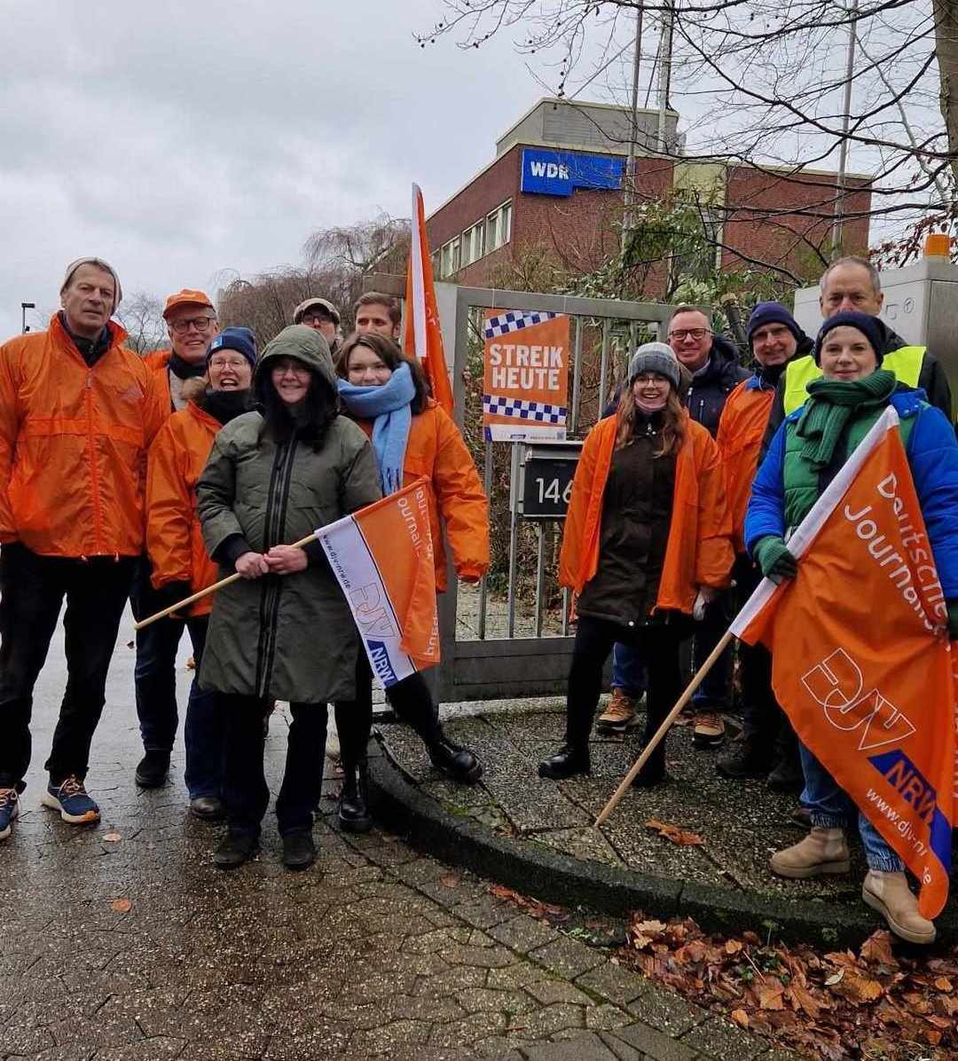 Viele Streikende in Münster in orangen Jacken und mit orangenen Fahnen. – Streikende Kolleg:innen trotzen vor dem WDR in Münster am 9.12.2024 Wind und Wetter.
